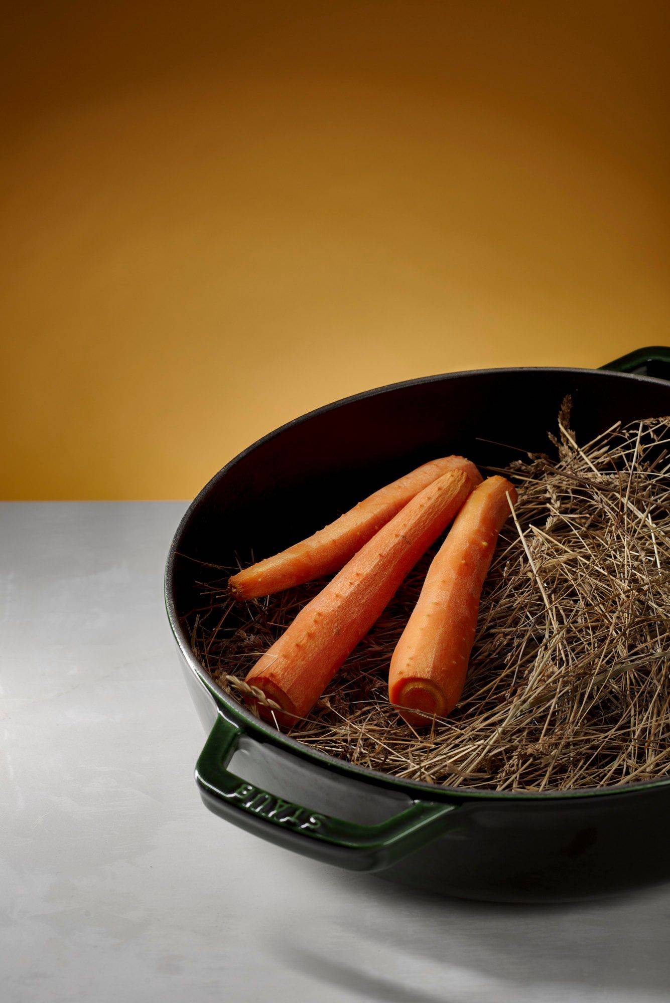 carrots in hay with green staub cast iron pot with white sapienstone top and yellow background