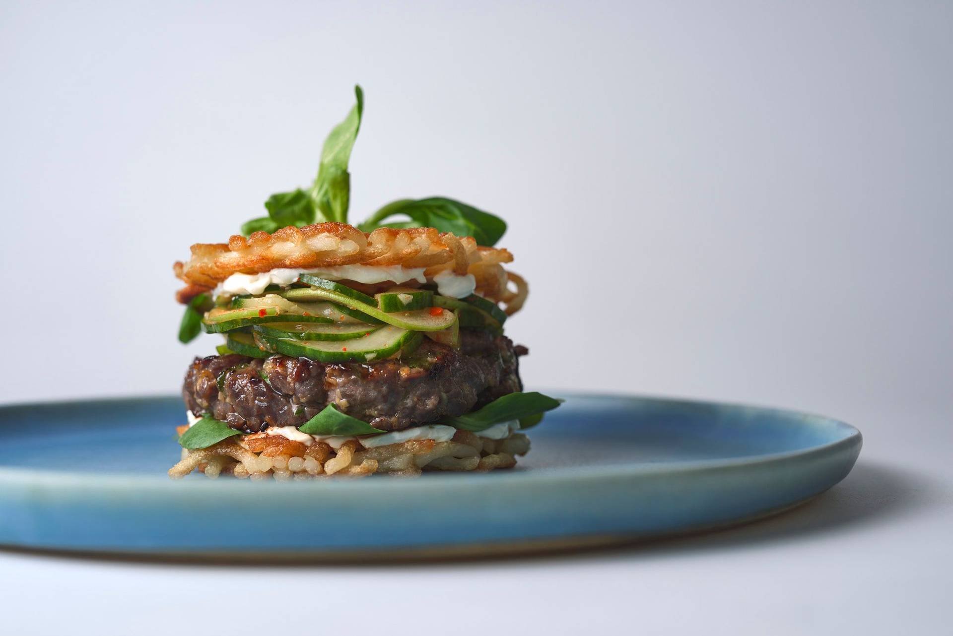 bulgogi beef burger with cucumber kimchi and udon noodle buns on a blue ceramic plate with white background