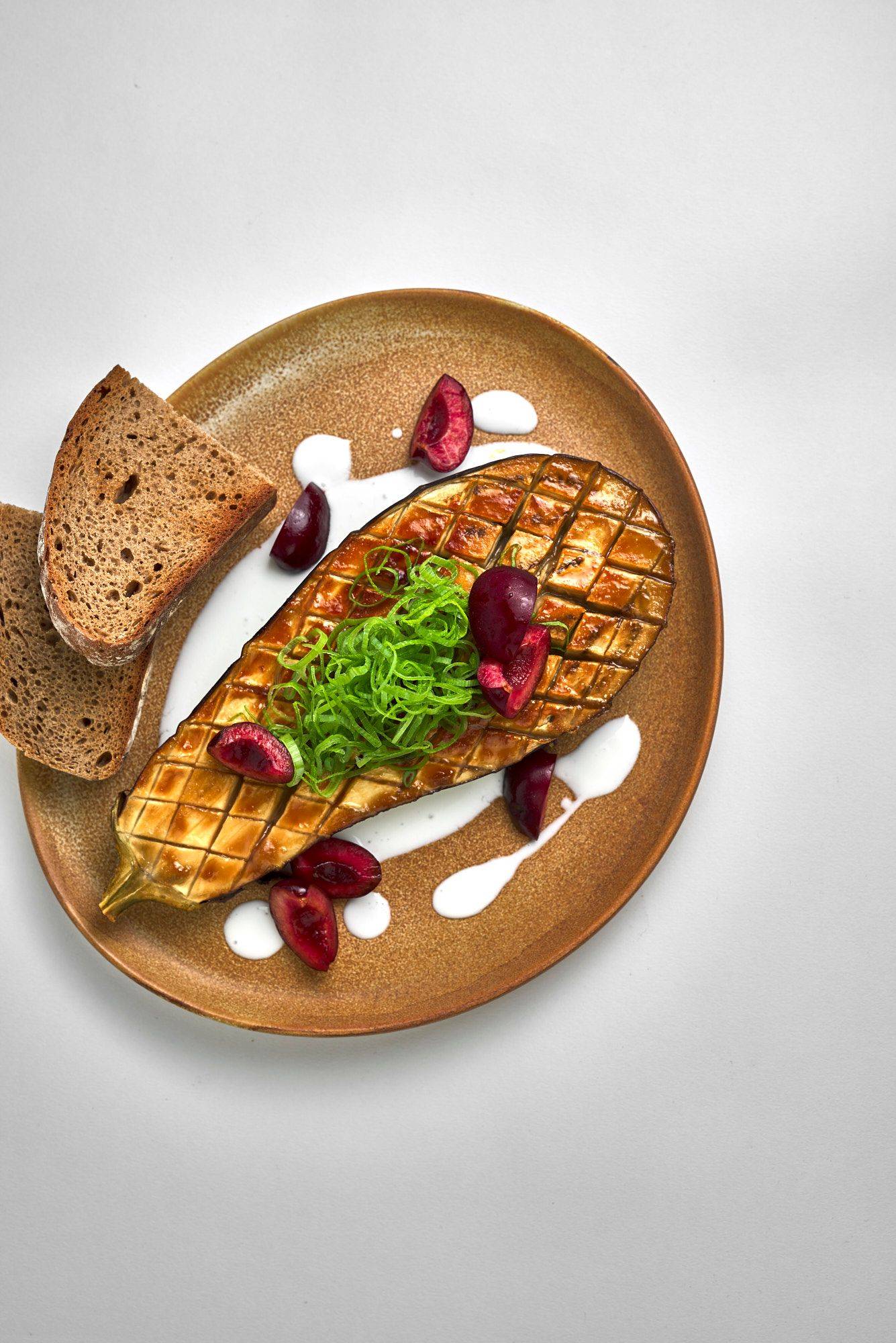 miso gebackene aubergine mit kefir kirschen und sauerteigbrot auf einem braunen keramikteller mit weißem hintergrund