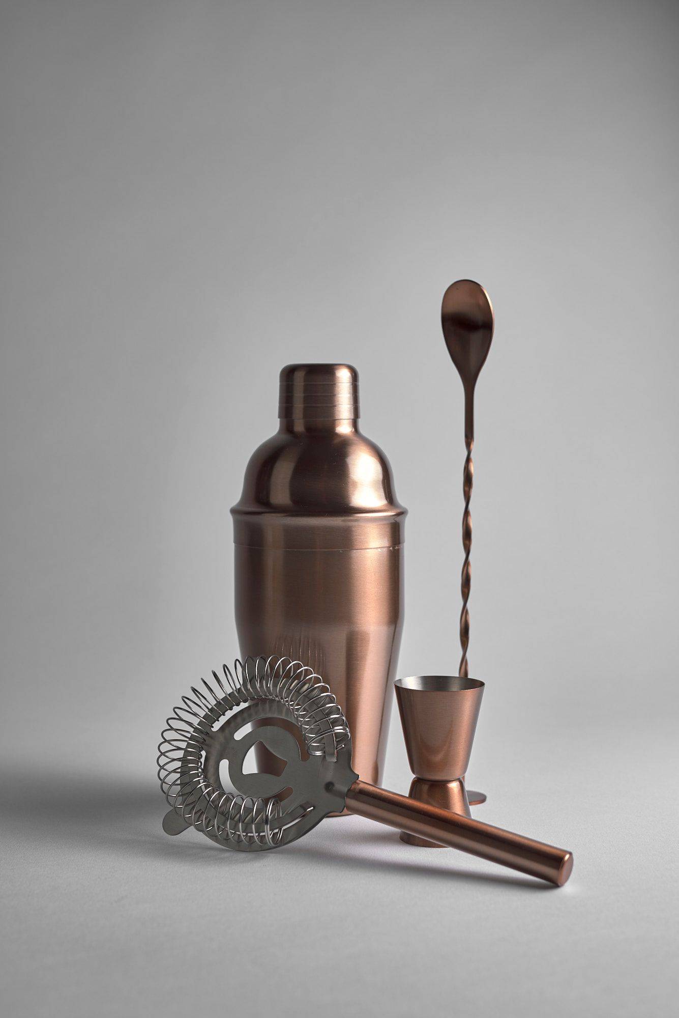 a copper jigger, spoon, strainer and measuring cup on white background 