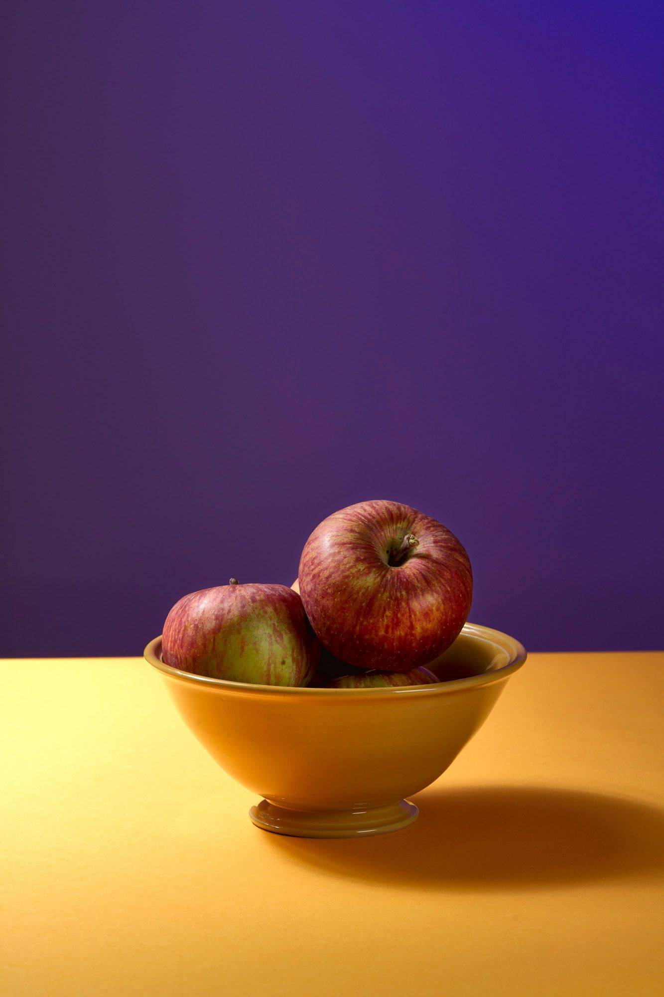 a bowl with three red apples with yellow and purple background