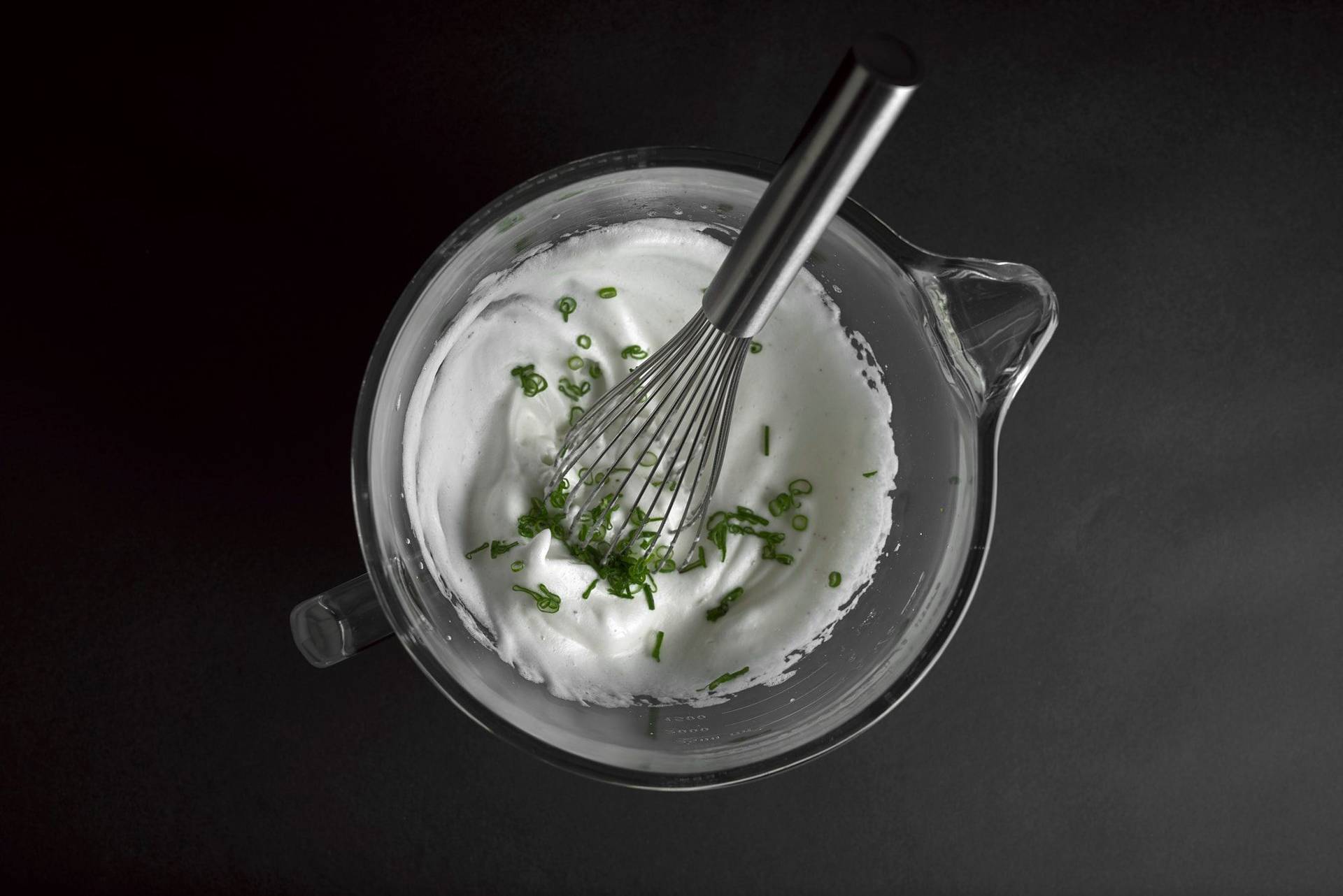 savory meringue with scallions in a glass mixing bowl with whisk on black background