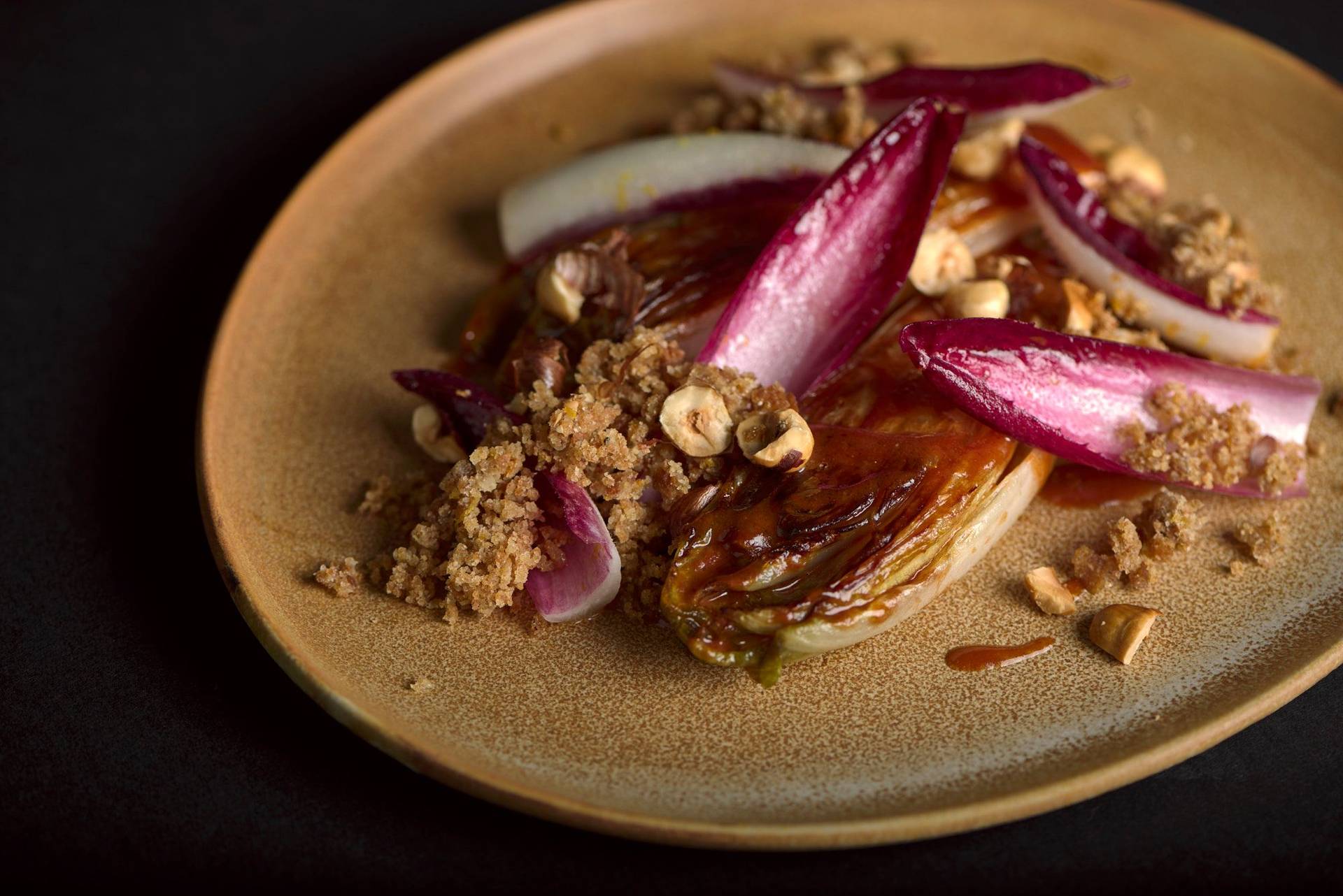 rosehip braised chicory with hazelnuts and crispbread on a brown ceramic plate with black background