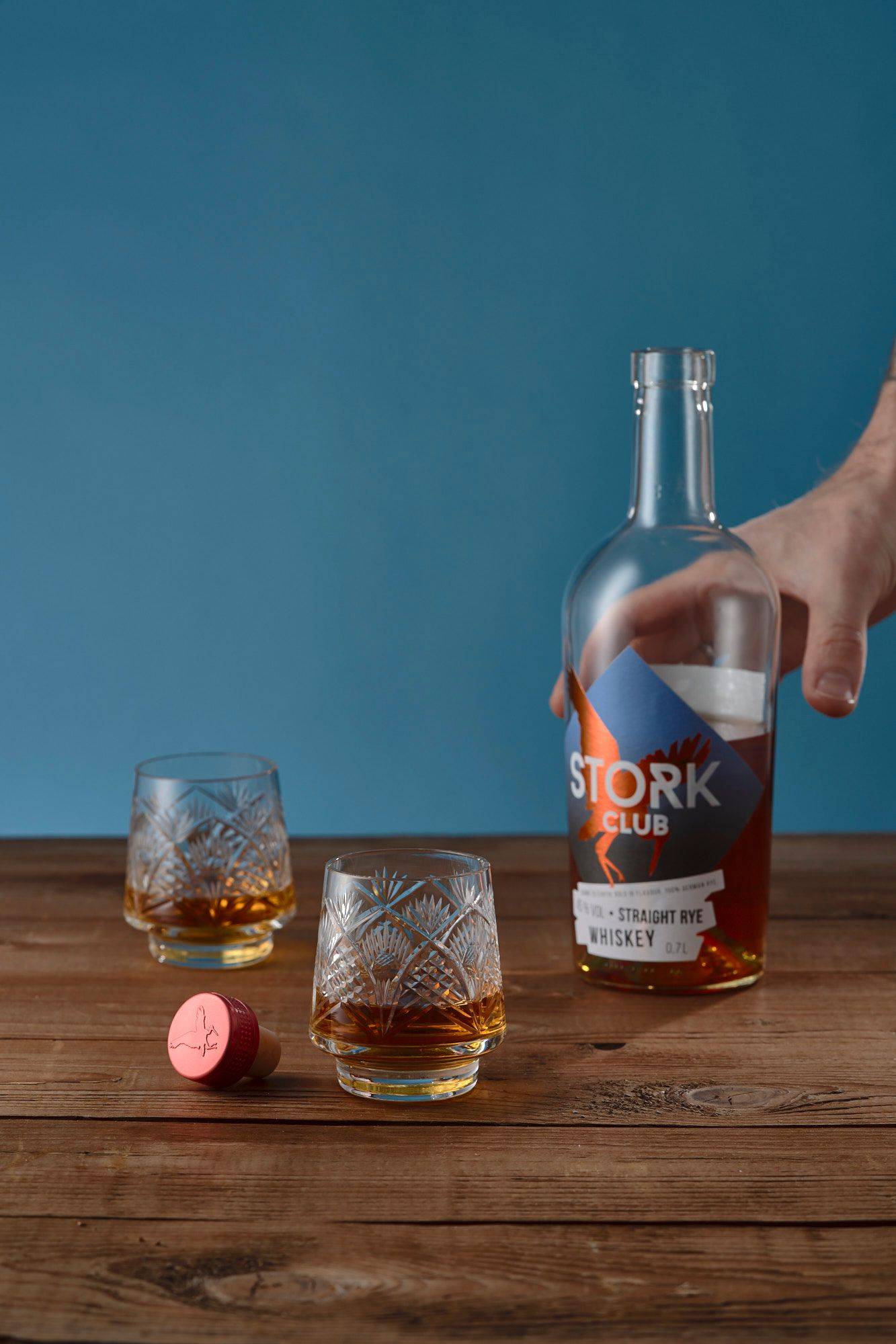 a bottle and two glasses with stork club german rye whiskey on a wooden table with blue background