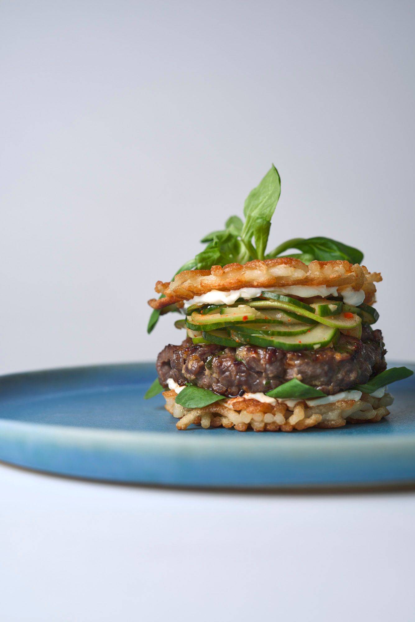 bulgogi beef burger with cucumber kimchi and udon noodle buns on a blue ceramic plate with white background