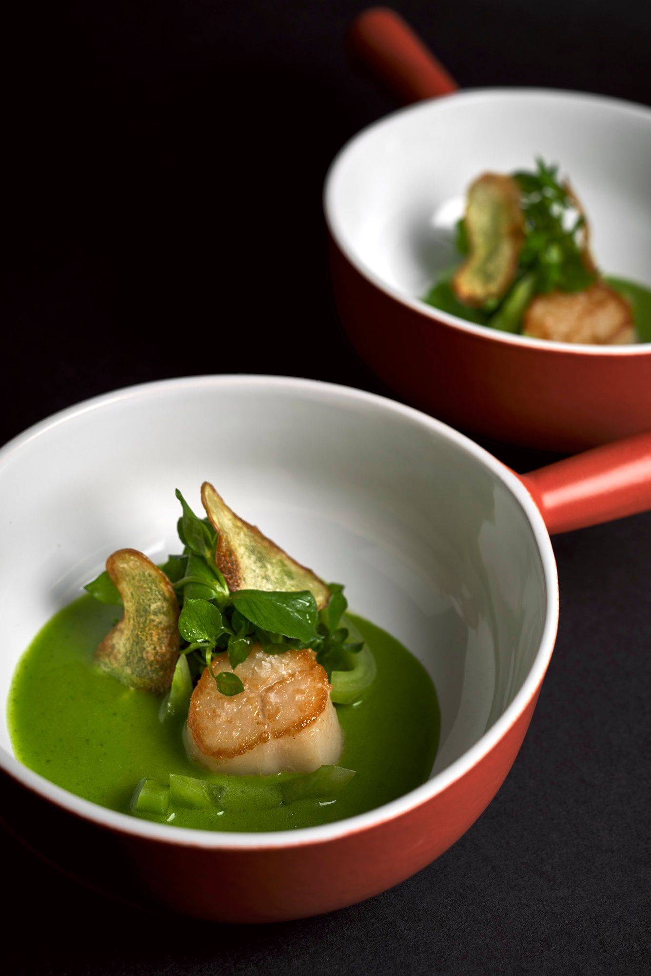two bowls of seared scallop with potato, wild garlic and sorrel sauce and chickweed on black background