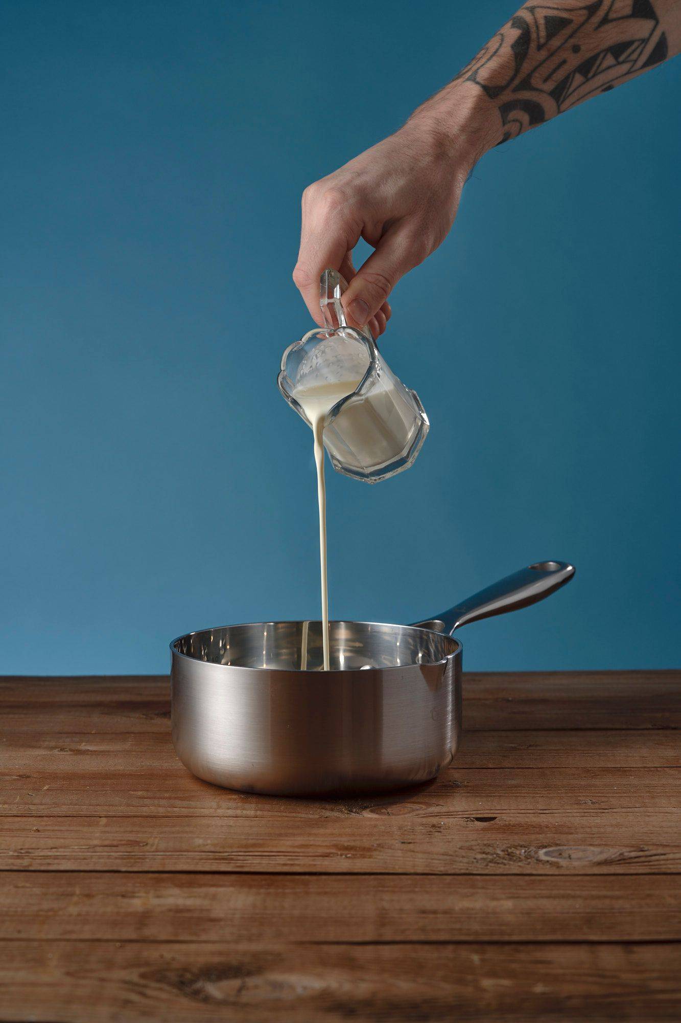 cream hand pouring cream into metal sauce pan on a wooden table with blue background