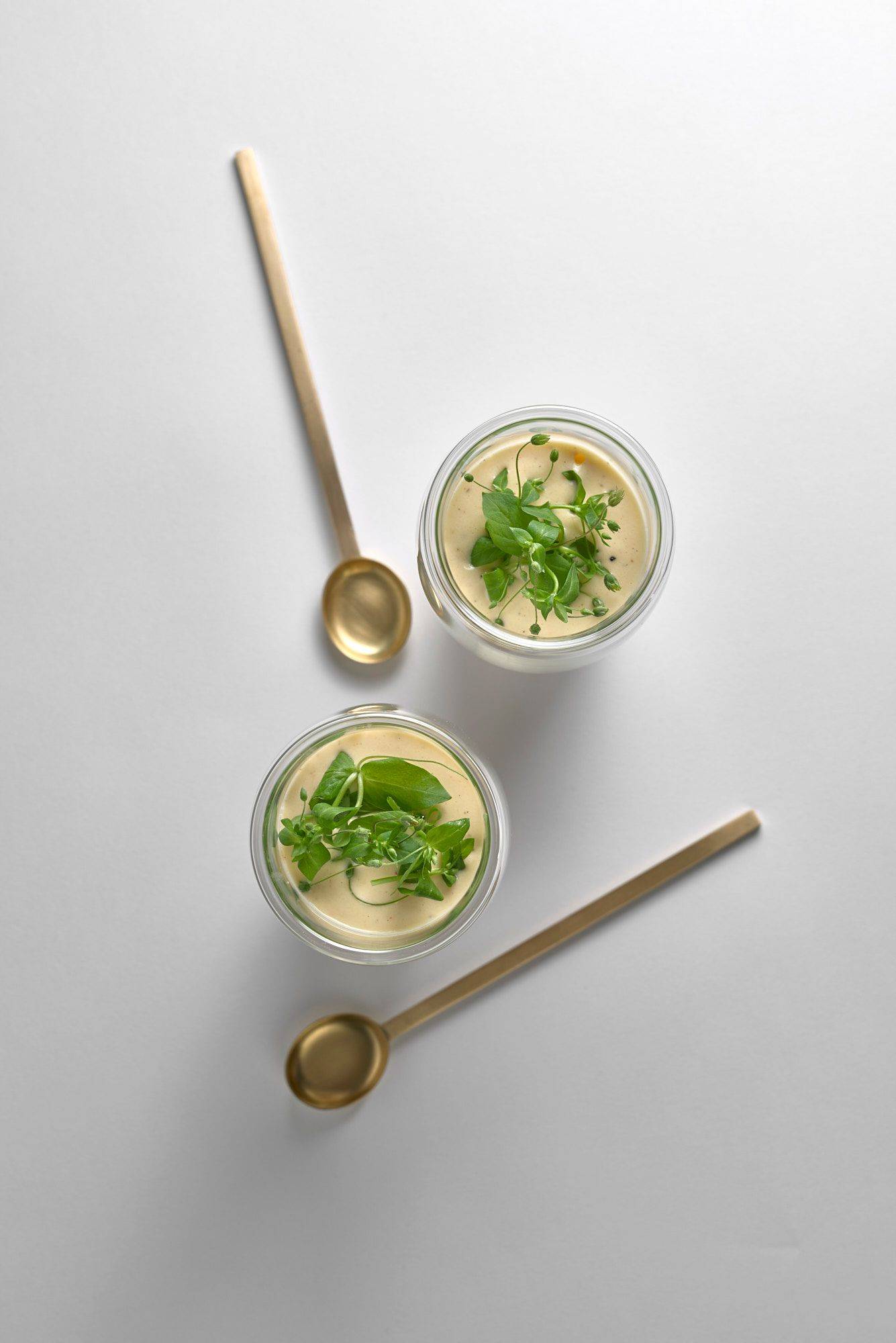 two glasses of savory asparagus cappuccino with brass spoons on white background