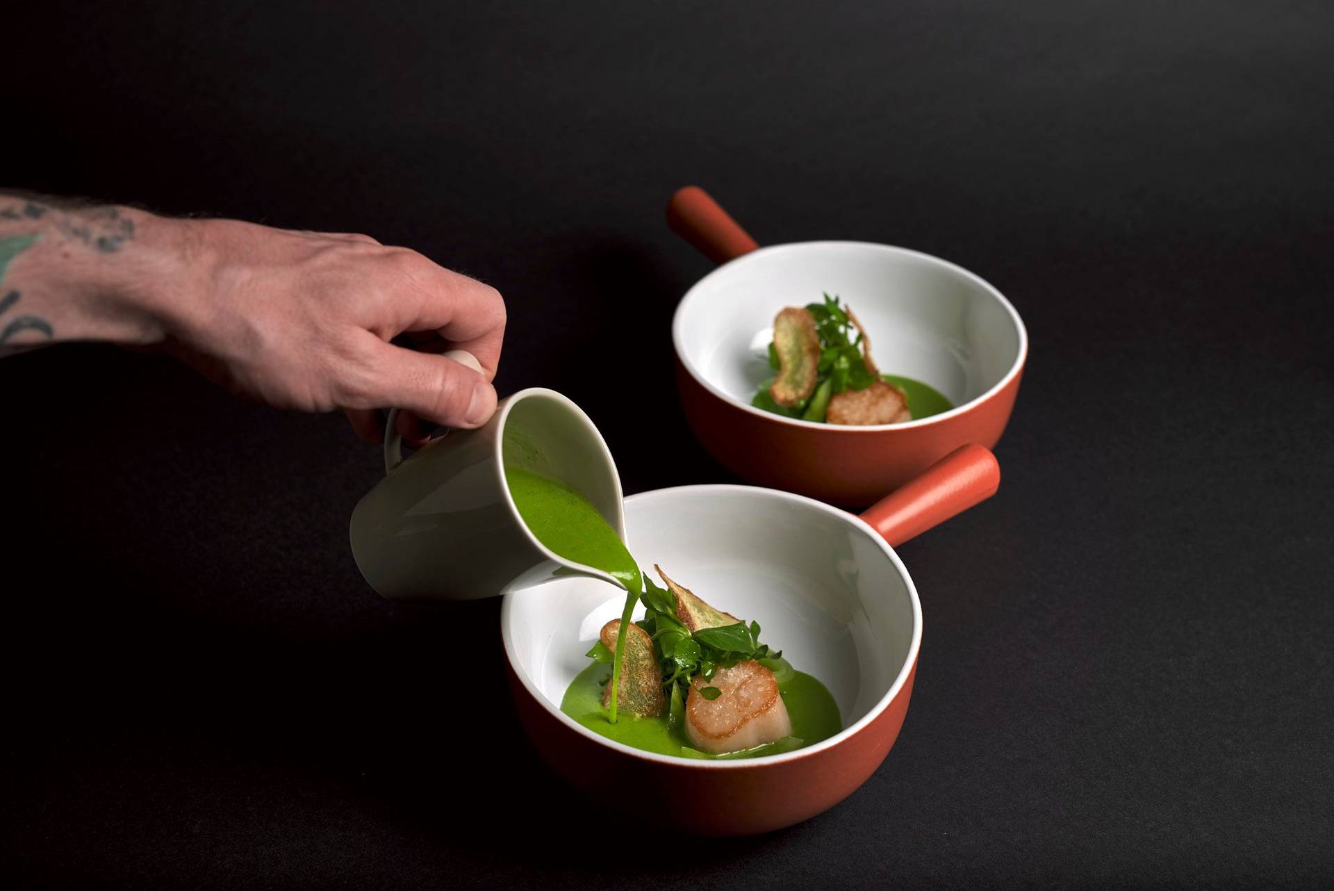 two bowls of seared scallop with potato, wild garlic and sorrel sauce and chickweed on black background