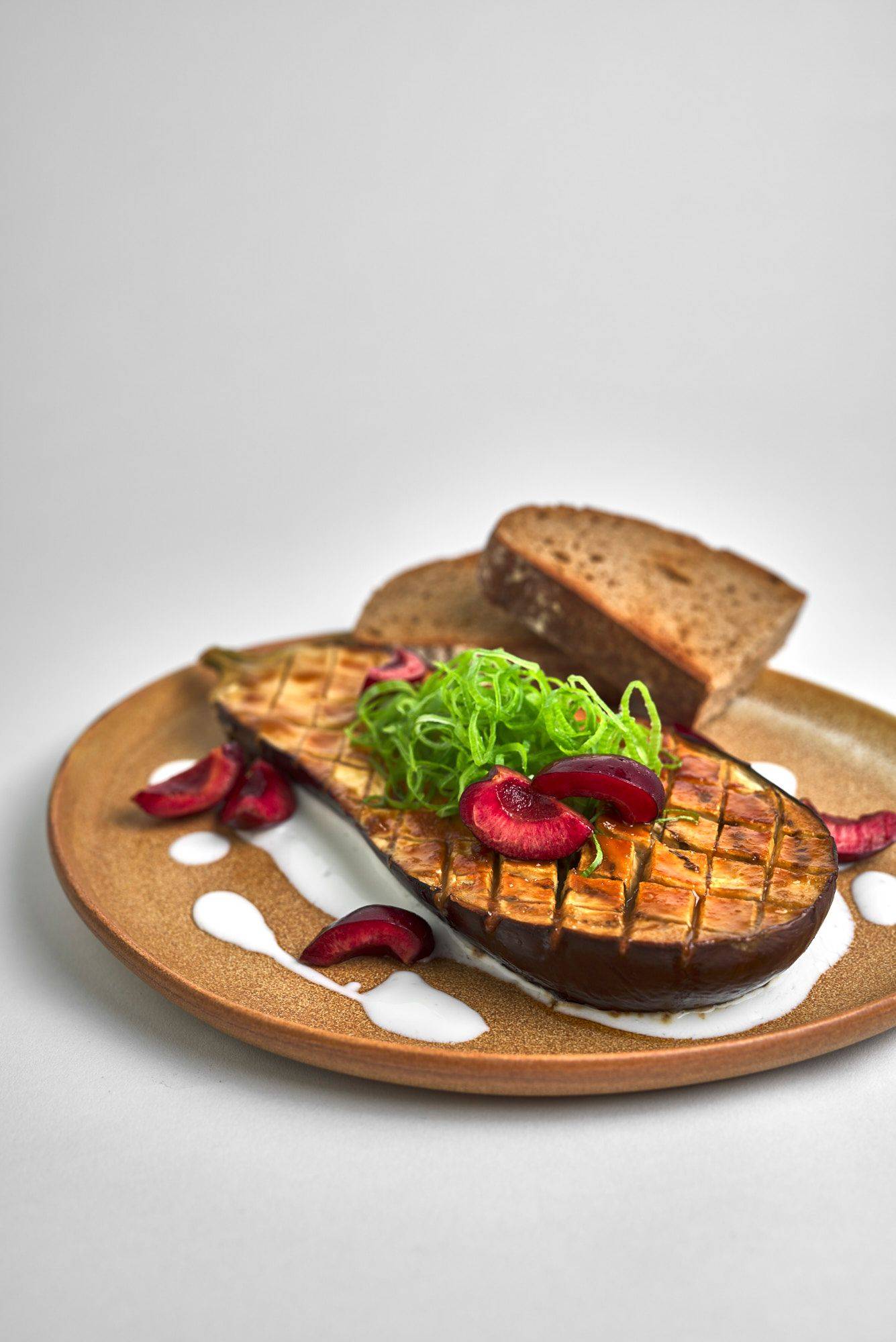 miso roasted eggplant with kefir cherries and sourdough bread on a brown ceramic plate with white background