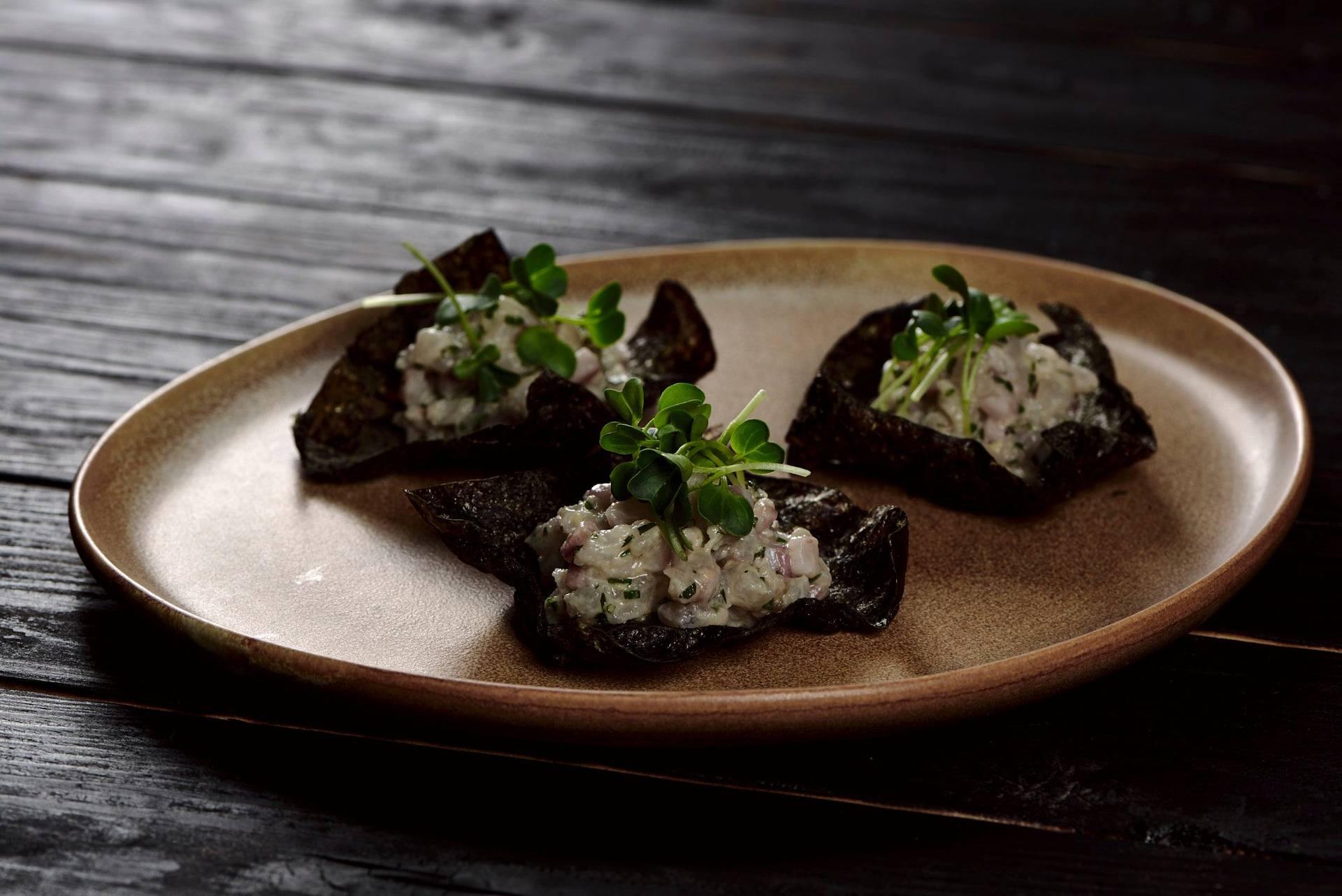 wolfsbarsch tatar mit nori cracker auf einem braunen keramikteller mit schwarzem holzuntergrund