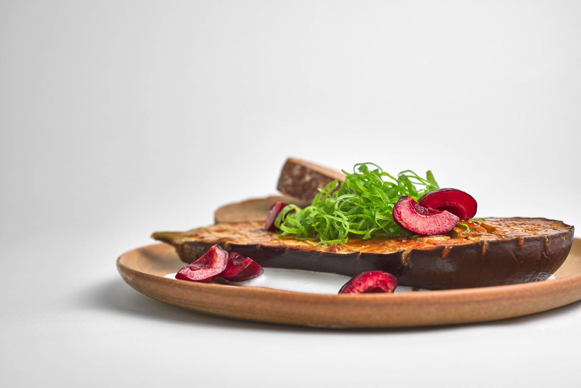 miso roasted eggplant with kefir cherries and sourdough bread on a brown ceramic plate with white background