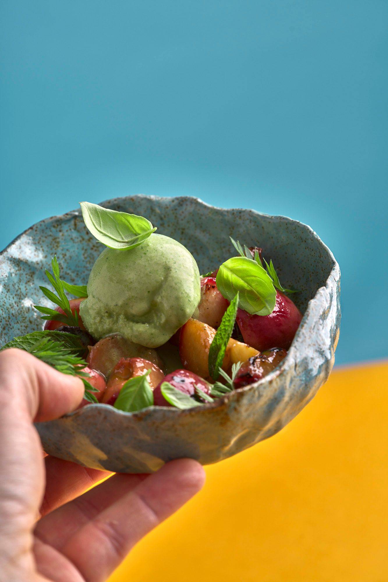 herbs and yogurt ice cream with grilled peaches and greek olive oil in a turquoise ceramic bowl with yellow and blue background