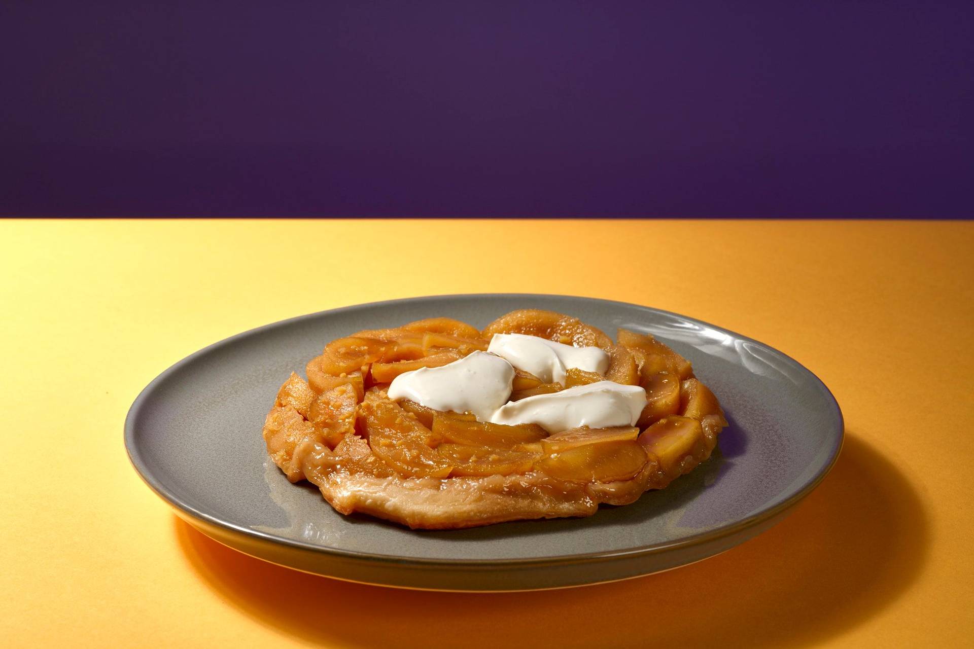 apfel und ingwer tarte tatin mit creme fraiche auf einem grauen teller mit gelbem und lila hintergrund