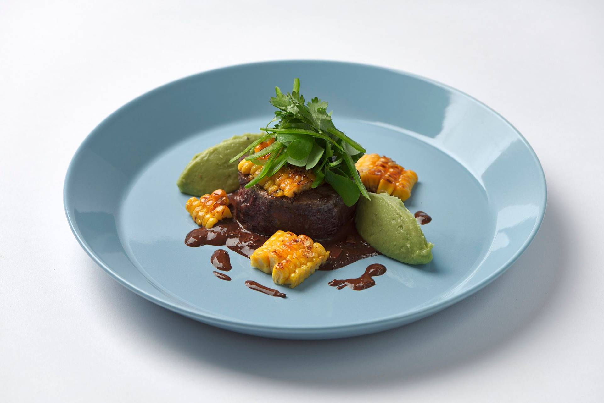 rinderfilet mit schwarzbier mole sauce mais und guacamole auf einem blauen teller mit weißem hintergrund